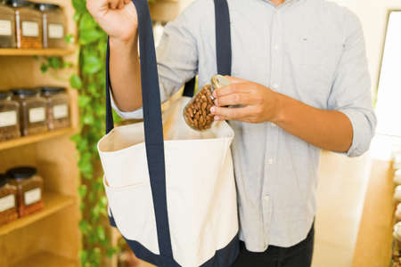 Latin young man putting a glass container filled with almonds into his shopping bag at the zero-waste supermarketの写真素材