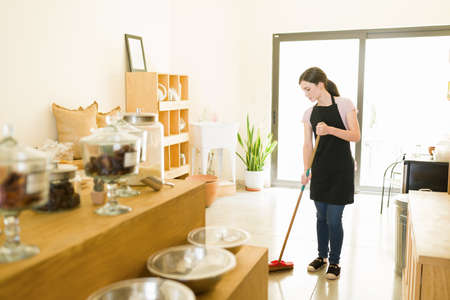 Hardworking female worker with an apron sweeping and cleaning the floors of a sustainable and...