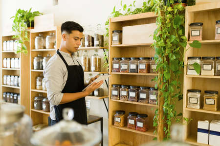 Young man working at a zero-waste bulk shop. Male worker holding a tablet and checking the prices of...