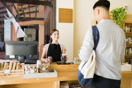 Beautiful young woman smiling and talking to a customer while working at the eco-friendly bulk shop. Customer paying at the marketの写真素材