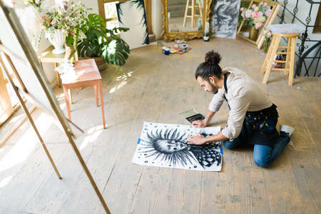 Talented young man sitting on the floor and drawing a portrait with charcoal technique at the studioの写真素材