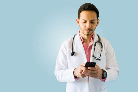 Attractive serious doctor with a lab coat and stethoscope texting his patients on the smartphone