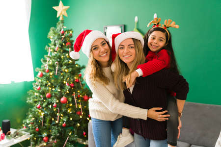 Happy  women and moms with a santa hat piggybacking her cute daughter. Multiracial family hugging...