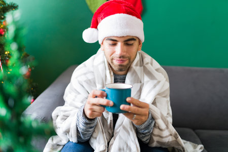 Happy handsome man wrapped in a blanket during a cold Christmas morning while drinking hot chocolateの写真素材
