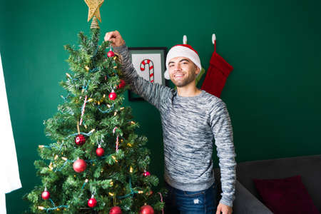 Portrait of an attractive young man decorating his Christmas tree and putting the star on topの写真素材