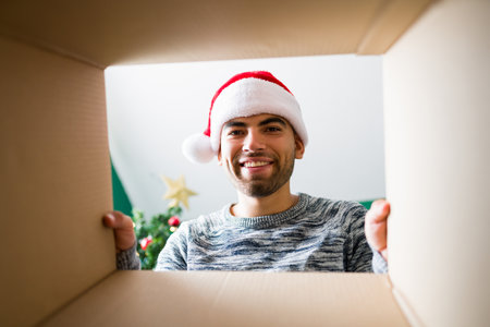 I love christmas gifts. Attractive young man opening a Christmas present and looking inside while...