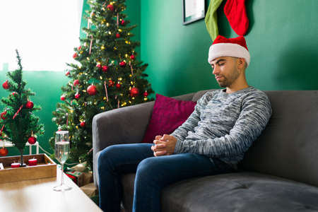 I feel so depressed. Sad hispanic man sitting alone at home and feeling lonely during Christmas eveの写真素材