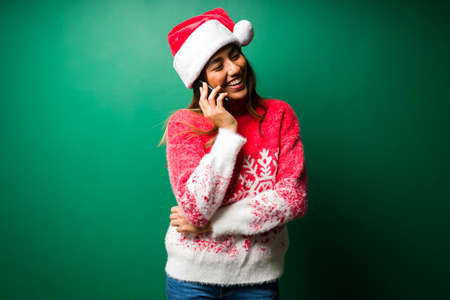 Cheerful woman talking on the phone and telling merry christmas to family and friends against a studio backgroundの写真素材