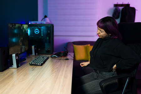 Tired young woman with a bad posture suffering from back pain while playing online video games on the computerの写真素材
