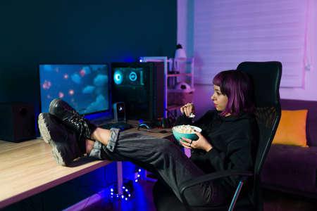 Beautiful young woman with purple dyed hair watching a movie or video game streaming on her computer while eating popcornの写真素材