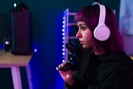 Profile of a beautiful latin gamer with dyed purple hair and headphones holding a gamepad while playing video gamesの写真素材