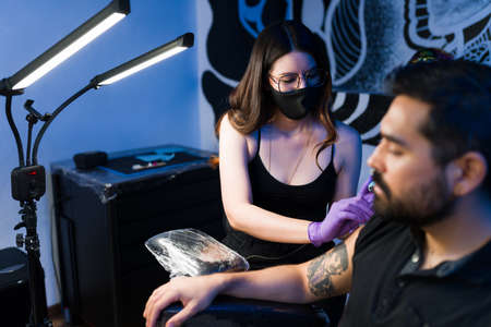 Beautiful tattoo artist with a face mask working on the tattoo of a male customer at the ink studioの写真素材