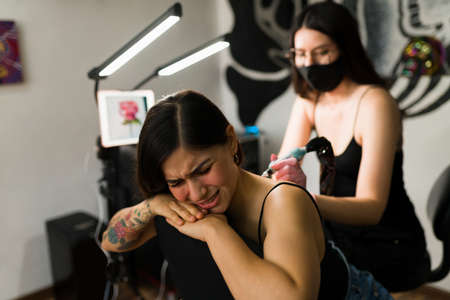 It hurts a lot. Stressed young woman getting a back tattoo at the ink studio with a professional female tattoo artistの写真素材