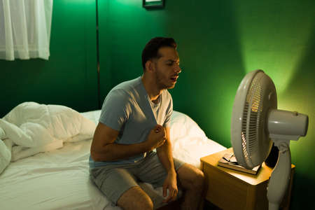 Distressed latin man sitting in front of the ventilator in bed and can't sleep because of the heat
