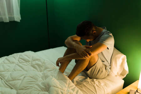 Depressed hispanic man in pajamas hugging his knees and feeling lonely in bed at midnightの写真素材