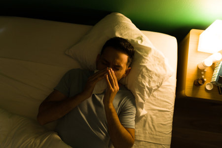 Feeling very sick. Hispanic man with a cold and fever blowing his nose while lying in bed at nightの写真素材