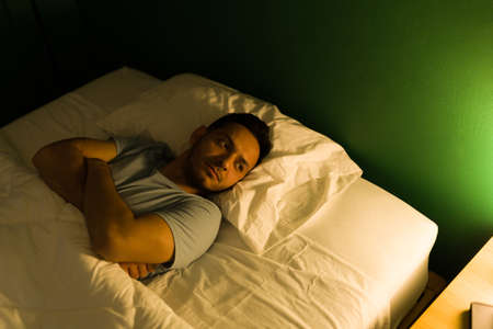 Insomnia concept. Tired and depressed man having trouble sleeping while thinking about his problems at midnightの写真素材