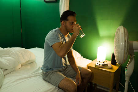 Hot night. Thirsty young man drinking water in front of a fan while sitting up in bed