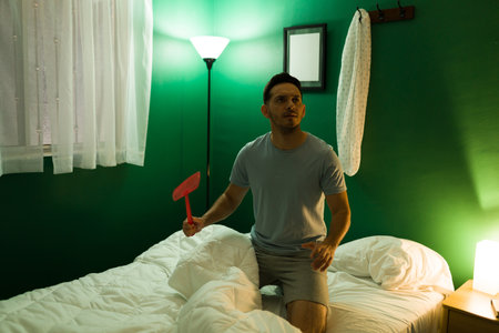 Upset young man trying to kill a fly in his bedroom because it won't let him sleep at nightの写真素材