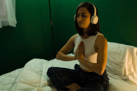 Prayer position. Calm young woman doing a meditation routine while listening to relaxing music with headphones in bedの写真素材