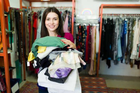 Gorgeous young woman shopping a lot of second hand clothes. Female customer about to pay vintage...