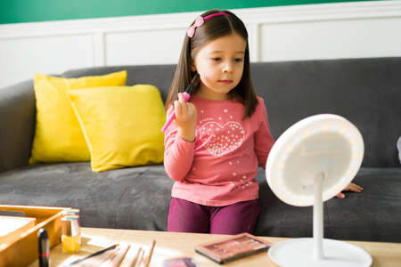 Adorable little girl having fun and playing. Cute kid looking in the mirror and using a make up brush to put on blush on her faceの写真素材