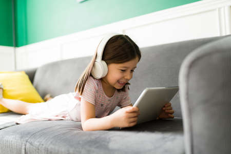 Excited elementary kid relaxing on the couch while watching online videos or movies on the tablet at homeの写真素材