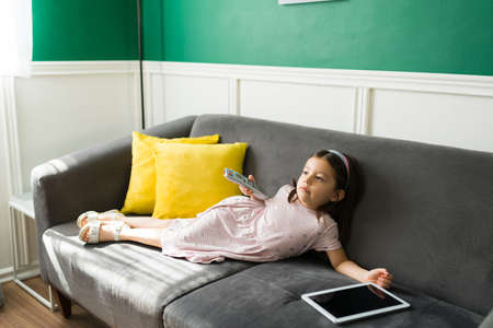 Relaxed little girl resting on the sofa and holding the tv remote while watching a movie and eating popcornの写真素材