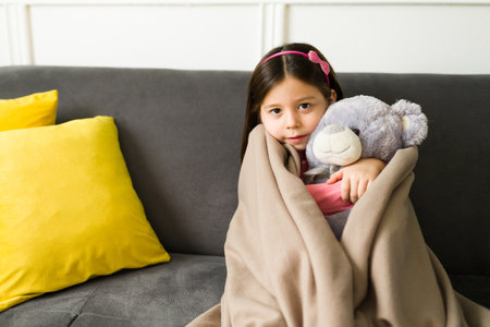 It's so chilly outside. Beautiful child hugging her teddy bear and feeling cozy under a blanket while relaxing during winterの写真素材