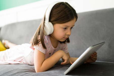 Beautiful 5 year old girl using a tablet and playing video games. Little kid with headphones having fun with online videos at homeの写真素材