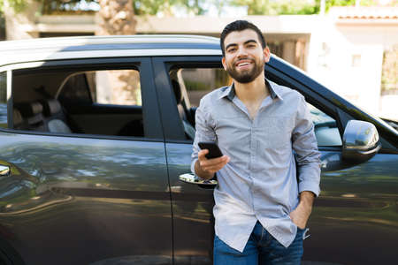 Smiling young man using his smartphone and texting while leaning on a black car outside