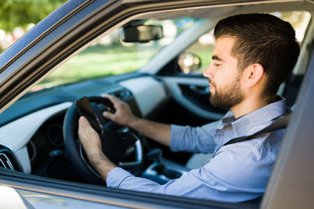 Happy young man accepting a new passenger ride on the rideshare app. Latin driver using his smartphone while drivingの写真素材
