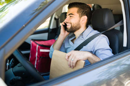 Cheerful delivery man calling a customer on the phone and delivering takeout food on his car