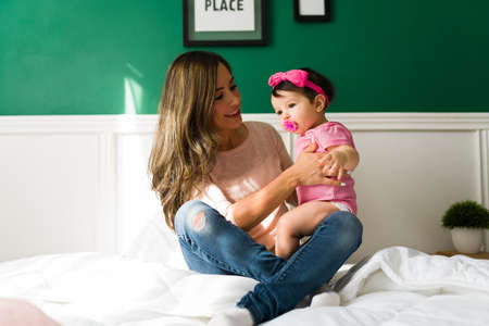 Attractive smiling mother putting her adorable baby girl in her lap while resting in bed together