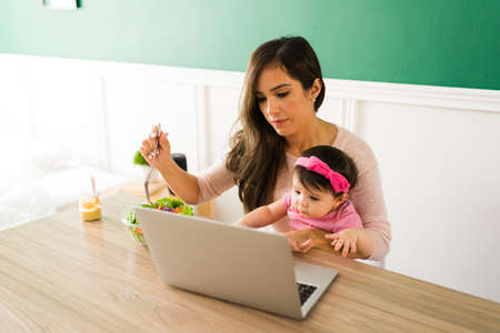 Busy young mother watching a video on the laptop while eating a bow of salad with her baby girl on...
