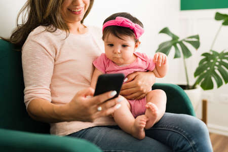 Caucasian young mom showing a video to her baby girl on the smartphone while sitting in the living...