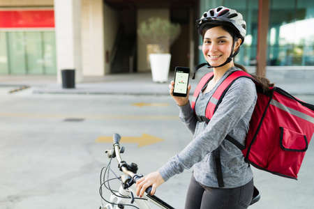 Cheerful delivery person smiling while showing her smartphone and using the GPS map of a food...