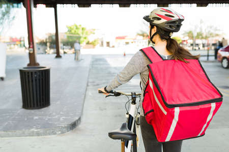 Delivery girl seen from behind with a red delivery backpack. Young woman riding a bicycle to make food deliveriesの写真素材