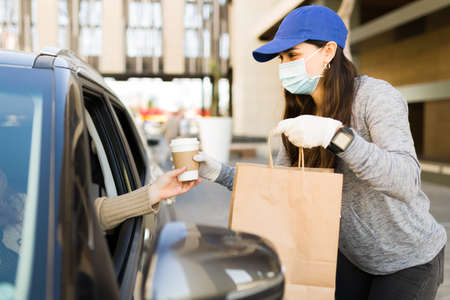 Hispanic delivery courier wearing a face mask and protective gloves while giving the food order to a customer in the carの写真素材