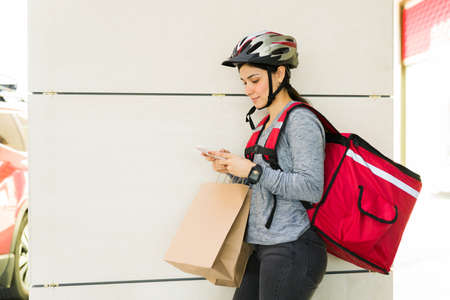 Happy delivery girl texting a customer to pick up the food order. Latin young woman waiting to make a deliveryの写真素材