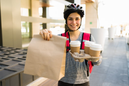 Making a food delivery. Smiling delivery girl making eye contact while holding a lunch bag and coffees to-goの写真素材