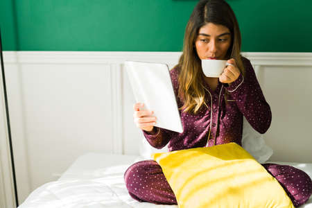 Reading online news. Hispanic woman looking at social media on the tablet while drinking coffee in bedの写真素材