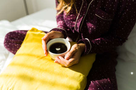 High angle of a woman in beautiful pajamas sitting in bed and enjoying a delicious cup of coffeeの写真素材