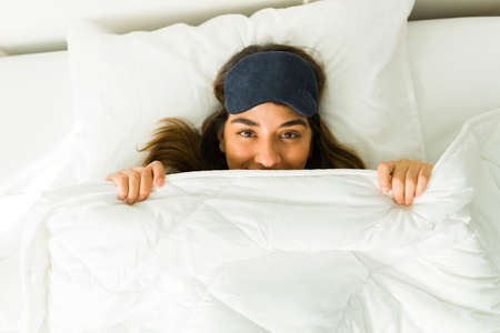 I love my bed. Happy young woman smiling under the covers in bed with a sleep maskの写真素材