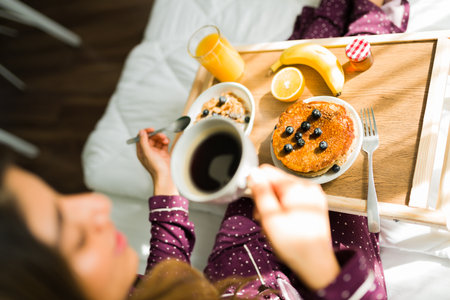 Top view of a young woman enjoying a delicious breakfast in bed. Tray with pancakes, blueberries, oats and coffeeの写真素材