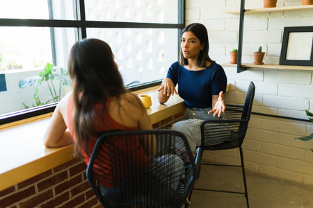 Latin young woman talking and catching up with a friend at a cafe while enjoying a cup of coffeeの写真素材