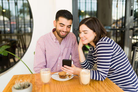 Happy woman using her smartphone and showing her boyfriend a text or online video on social media at the coffee shopの写真素材
