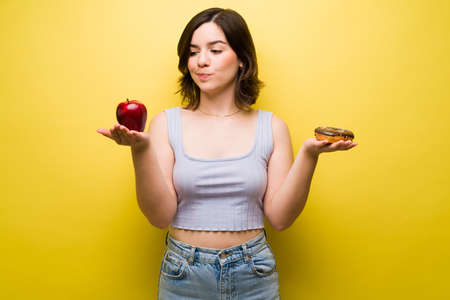 What should I eat? Hungry woman holding an apple and donut while deciding what to eat for a snackの写真素材