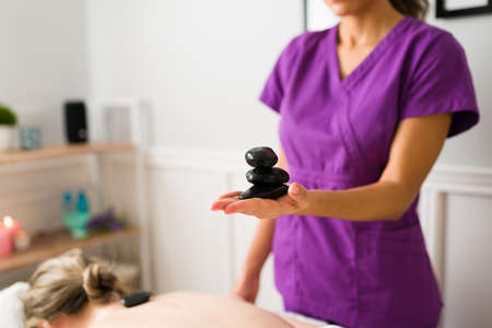 Relaxed spa treatment. Latin therapist holding hot stones to give a massage to a mature clientの写真素材