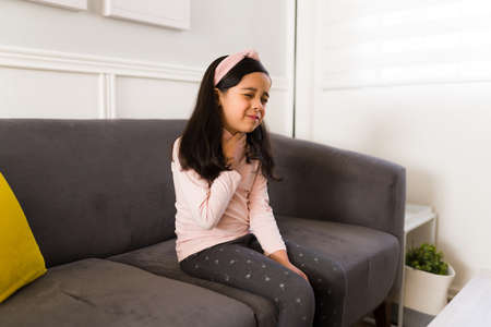 Sick elementary girl suffering from a sore throat and touching her neck while resting at home